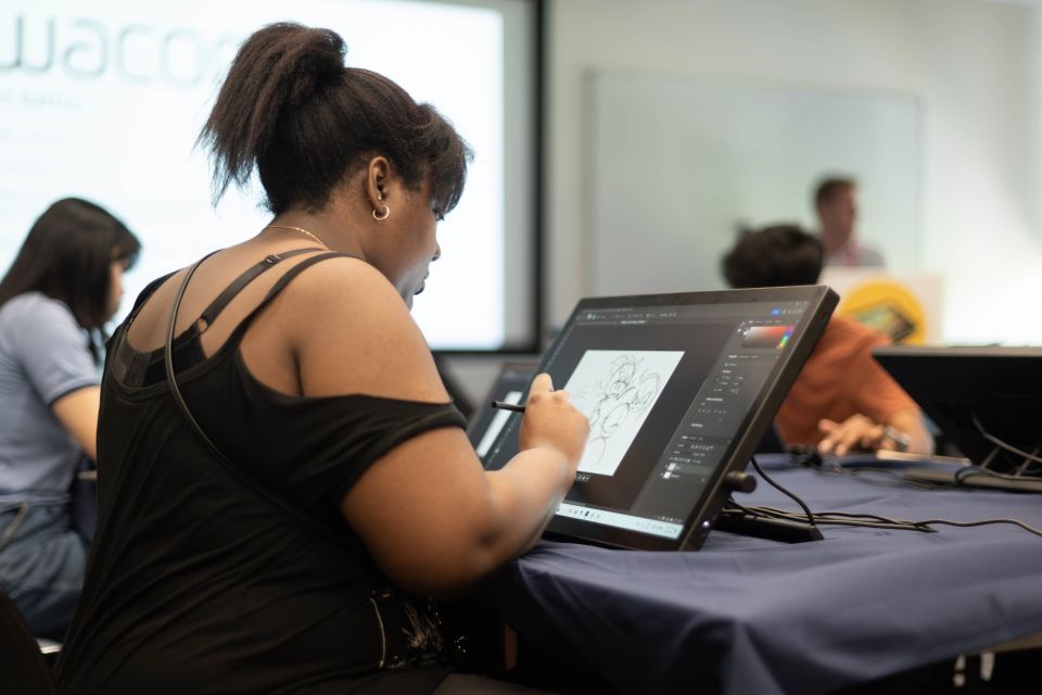 Ayoung  woman uses a stylus to draw on a digital tablet, with other people working in the background.