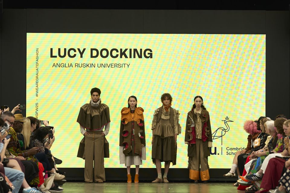Four models showcase earthy-toned outfits on a runway, with a large screen displaying "Lucy Docking, Anglia Ruskin University" in the background.