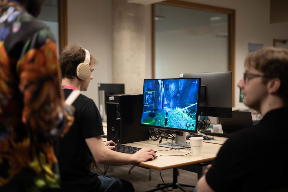 Young man wearing headphones, playing a video game on a computer with two other people nearby.