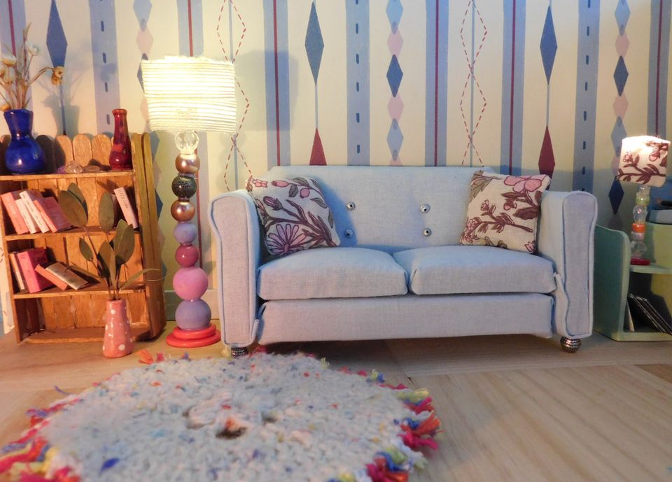 Photo of a lounge with grey sofa, grey mat with colourful fringes, lamp with stand made from coloured spheres, wooden bookshelf