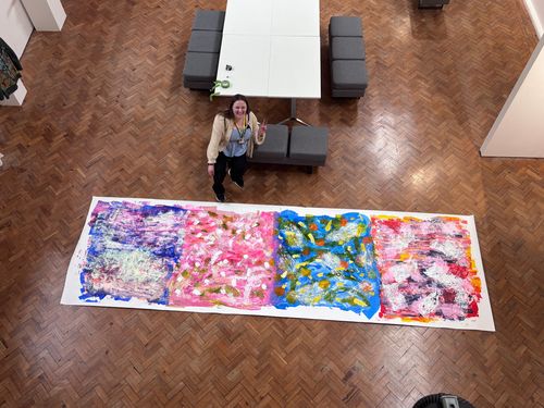 Woman standing next to a large, colorful abstract painting on a wooden floor, with a table and gray seats in the background.