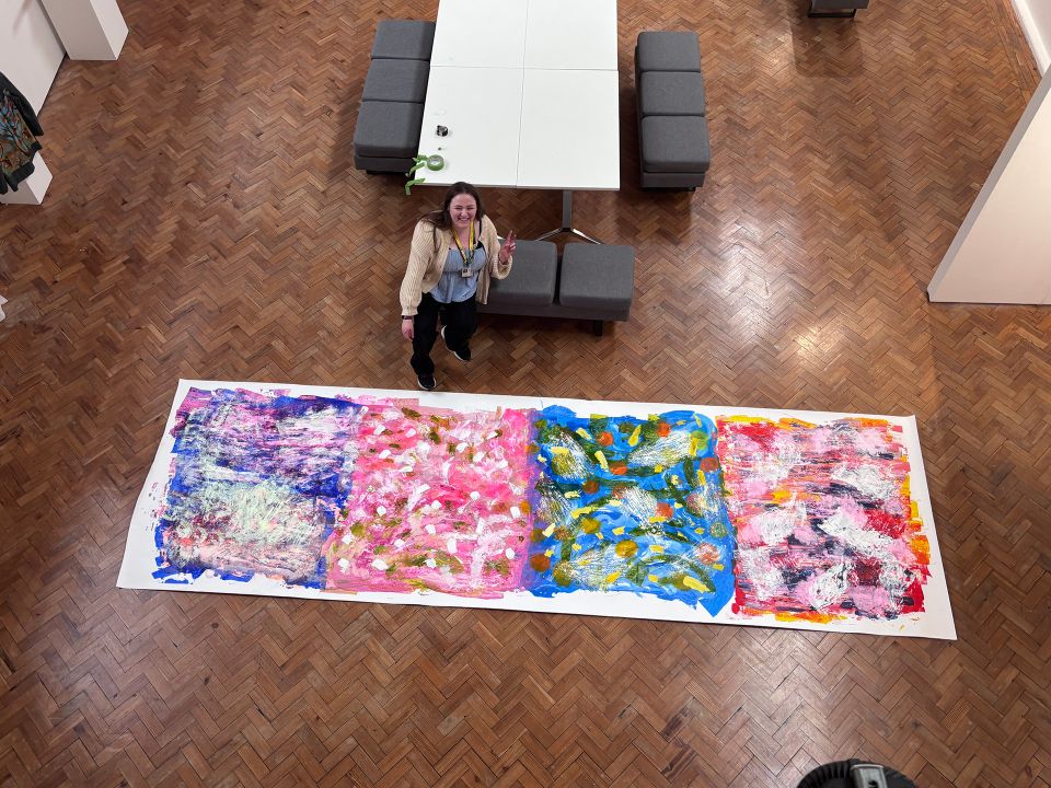 Woman standing next to a large, colorful abstract painting on a wooden floor, with a table and gray seats in the background.