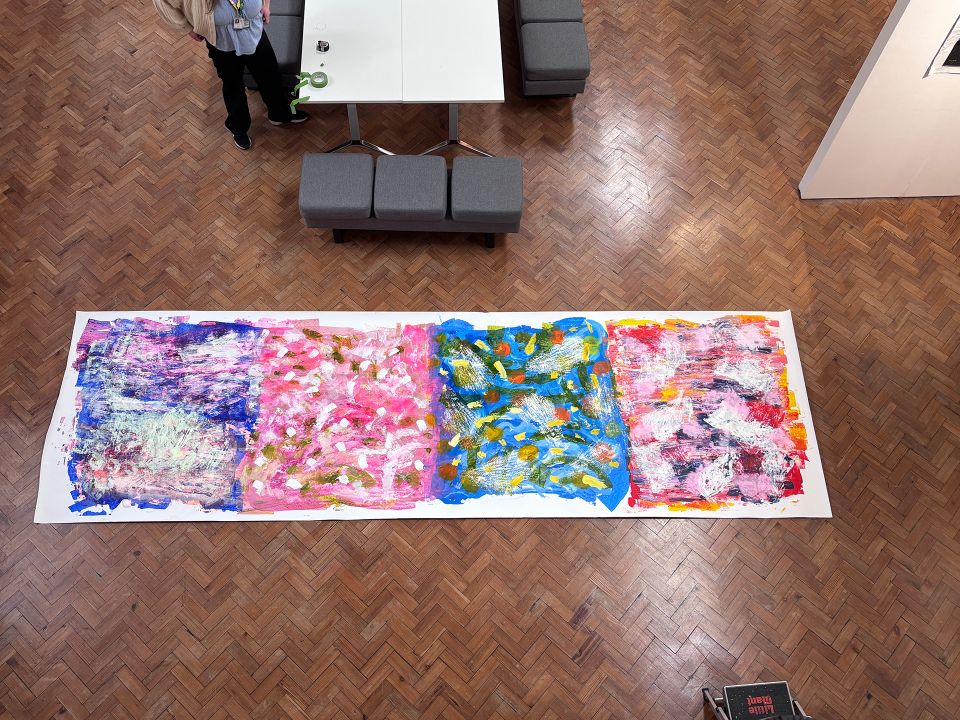 Colorful abstract painting on a large canvas on a wooden floor, viewed from above. A person stands nearby at a table.