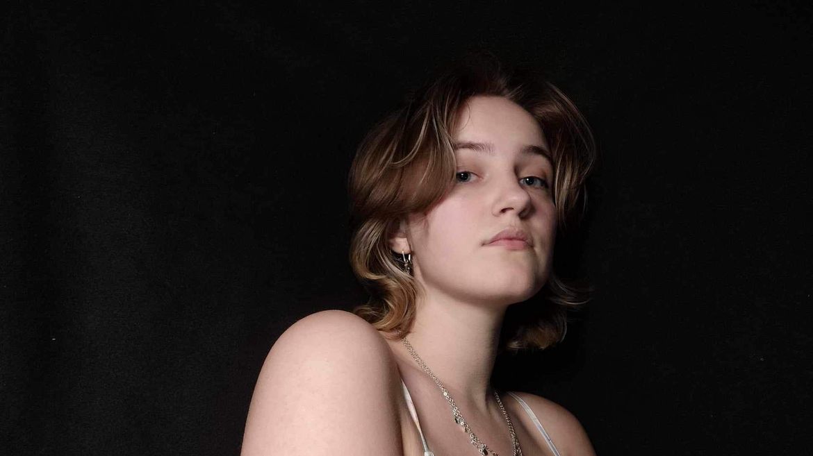 Maddie Schonningsen wearing silver necklace, bare-shouldered, gazing at camera against a black background.