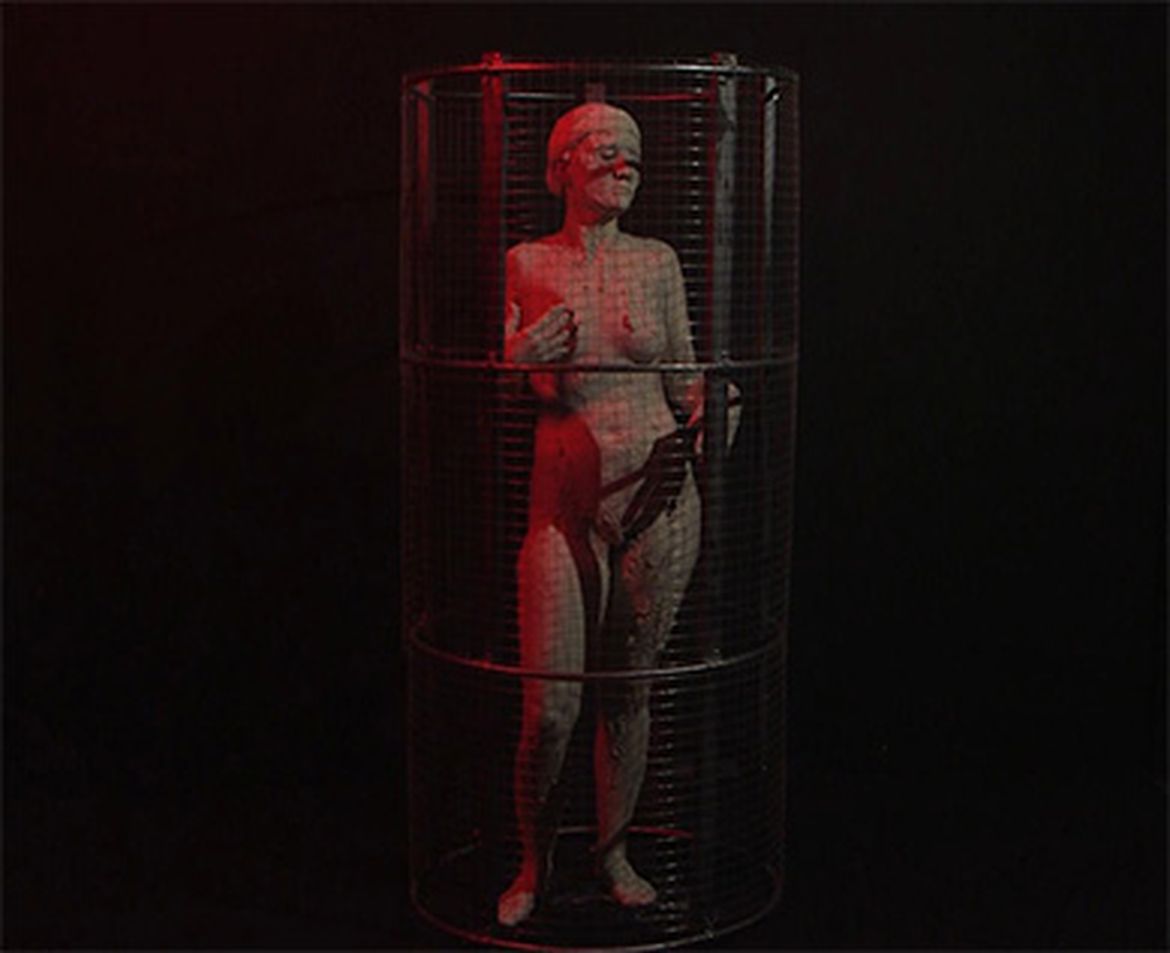 A sculpture of a person enclosed in a cylindrical wire cage, illuminated by red light against a dark background.