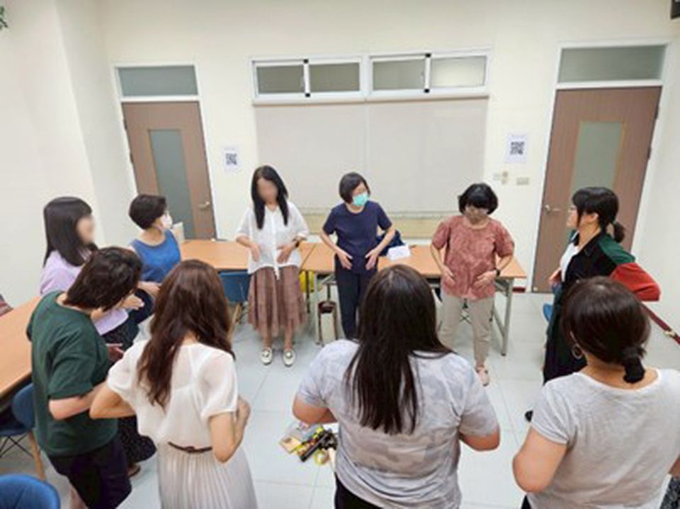 A group of people stand in a circle in a room, engaging in discussion. Some are wearing masks. Tables and chairs line the walls.