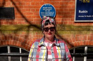 Gunta Andrews standing outside ARU's Ruskin building under a blue plaque for Syd Barrett