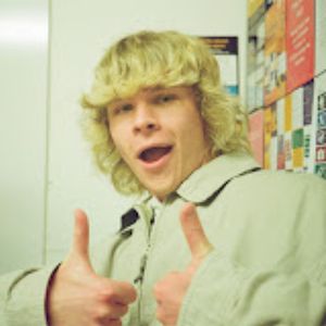 Jacob giving two thumbs up, smiling, wearing a light jacket, standing in front of a bulletin board.