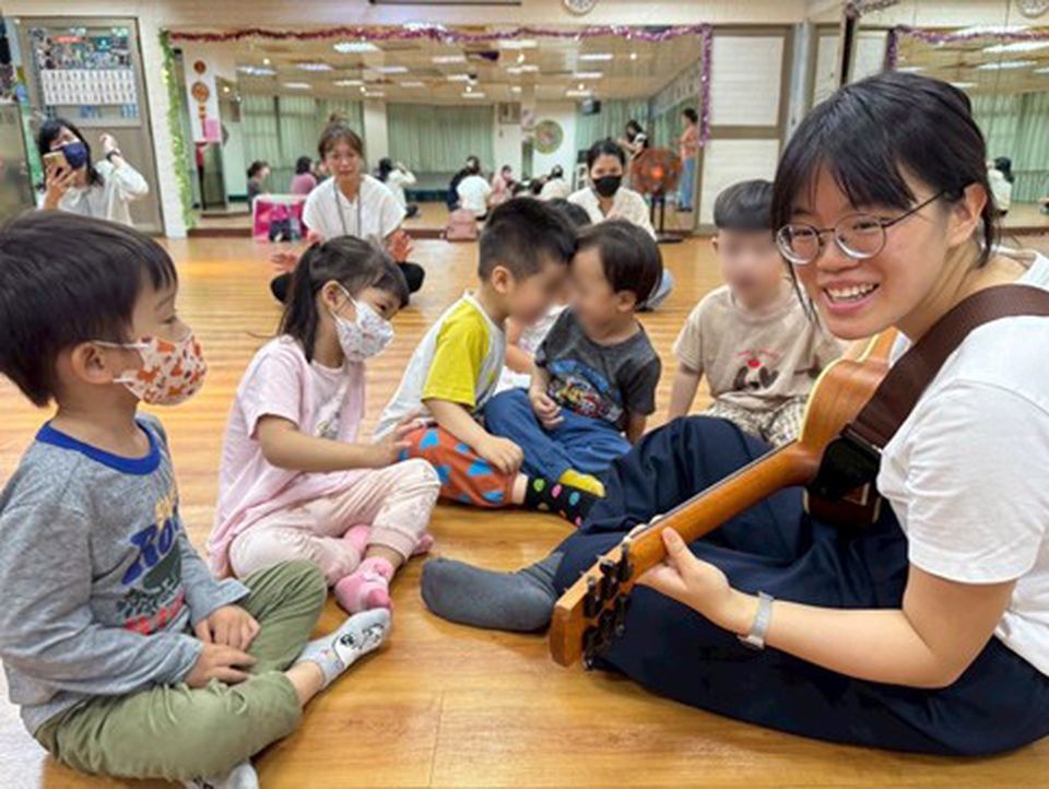 Amanda sits on the floor with a guitar, smiling at a group of children wearing masks, in a classroom setting.