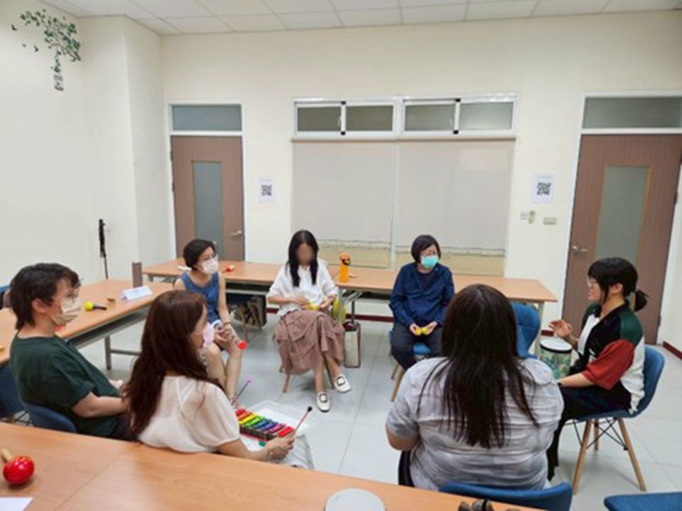 Seven people seated in a circle in a classroom, engaged in discussion. Some hold objects, and most wear masks.
