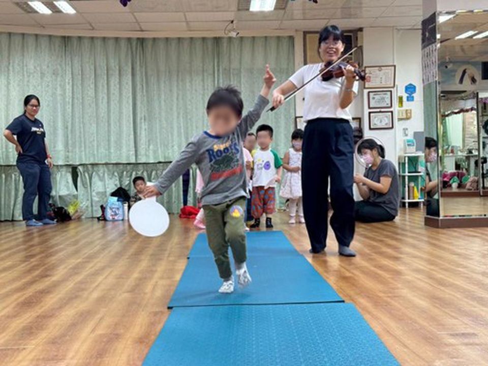 Child joyfully runs on a mat holding a tambourine, while Amanda plays the violin in a classroom with onlookers.