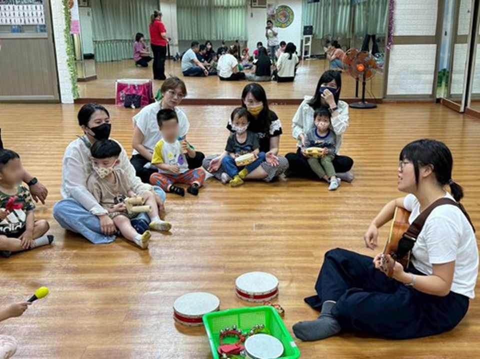 A group of adults and children sit on the floor in a circle, engaging in a music activity with instruments, while Amanda plays guitar.