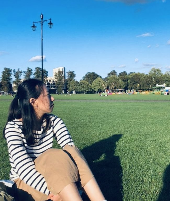 Charlee Tsai sitting on Parker's Peace, Cambridge, with clear blue sky, trees and a funfair in the background