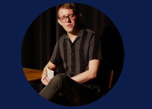 Jack Palmer sitting on stage on wooden chair, leaning on wooden table