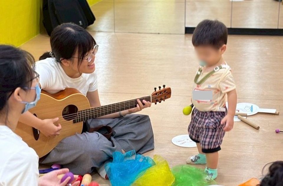 Amanda plays guitar for a toddler in a playroom, surrounded by colorful toys and scarves. Another adult is nearby, wearing a mask.
