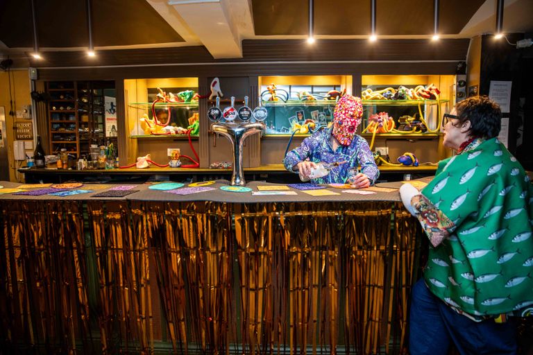 An image of a bar with a figure wearing a red mask embekllished with buttons and wearing a blue, white and green patterned top is serving a drink to a figurewearing a green top decorated with a tunafish pattern.