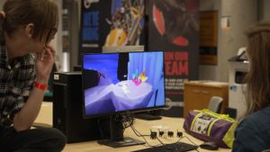 Two young women watching a computer screen displaying a colorful video game scene.