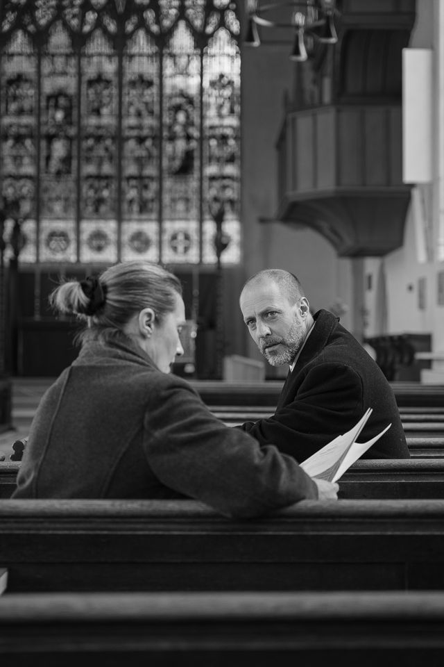 A man and woman sit on church pews, engaged in conversation, with one holding papers. Stained glass windows and ornate architecture are visible in the background.