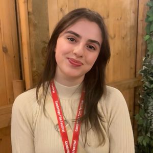Woman smiling, wearing a cream sweater and red lanyard with "Cambridge Film Festival," standing against a wooden background with greenery.