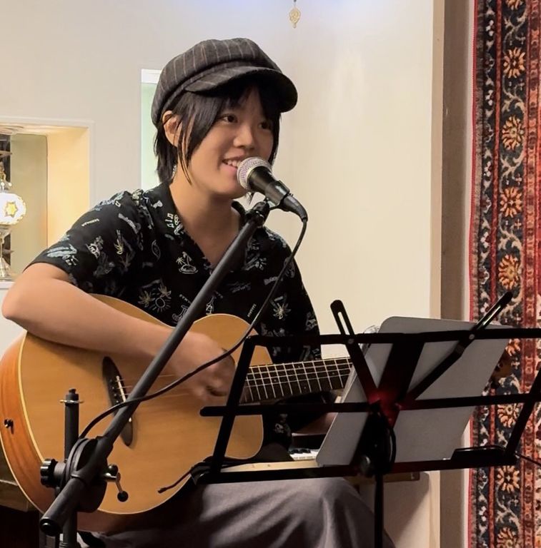 Amanda Lee playing an acoustic guitar and singing into a microphone, seated indoors with a music stand in front.