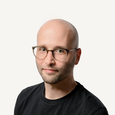 Portrait of Thomas Weber, a bald man with a short beard and round tortoiseshell glasses, wearing a black T-shirt and smiling slightly against a plain off-white background.