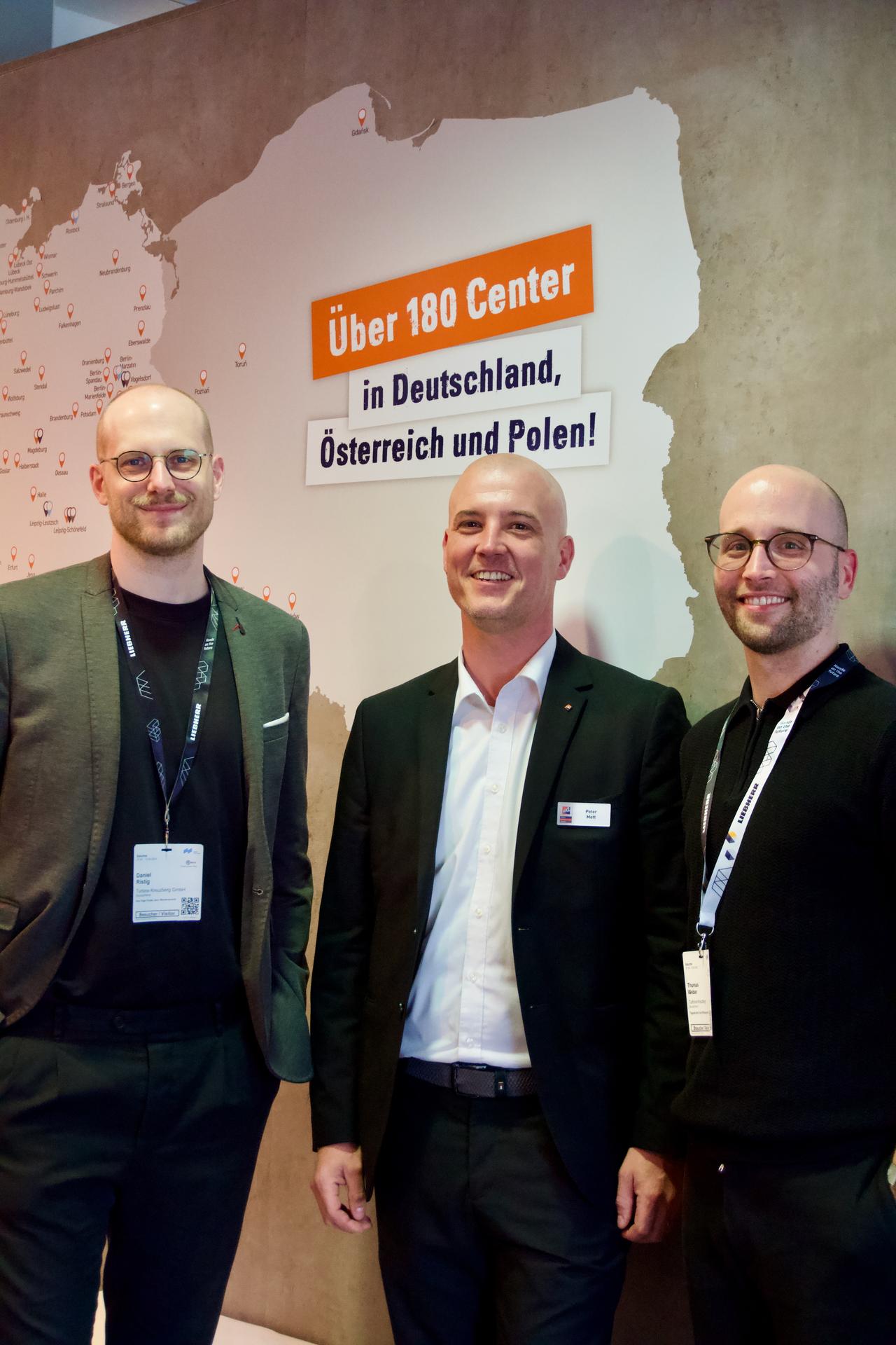 Three individuals standing in front of a wall display featuring a map and the text "Über 180 Center in Deutschland, Österreich und Polen!" written in bold lettering. The individuals are dressed in business attire, and the setting appears to be a professional event or conference.