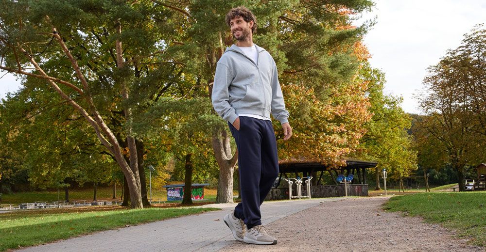 Ein Mann geht in Sportkleidung mit Sneakern einen von Bäumen gesäumten Parkweg entlang, lächelt und schaut zur Seite.