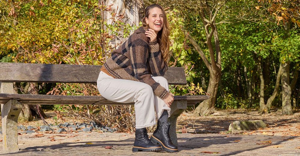 Eine Frau sitzt lächelnd auf einer Parkbank; sie trägt einen karierten Pullover, eine weiße Hose und schwarze Stiefel, im Hintergrund sind herbstliche Bäume zu sehen.