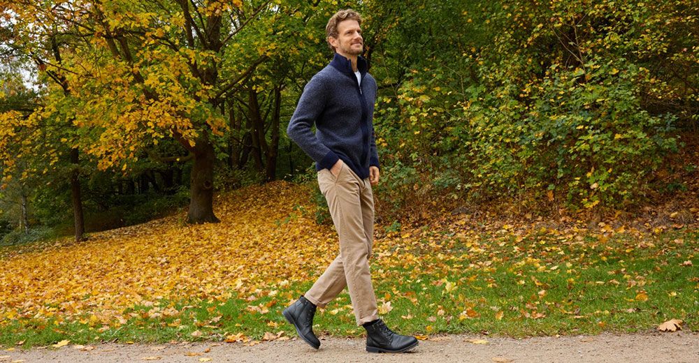 Ein Mann in einem blauen Pullover und einer beigen Hose geht auf einem mit Laub bedeckten Weg in einem herbstlichen Park spazieren, umgeben von Bäumen mit farbenprächtigem Laub.