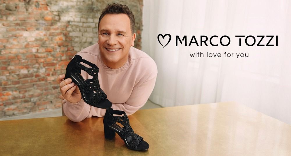 Eine lächelnder Mann (Guido Maria Kretschmer), der an einem Tisch einen schwarzen Riemchen-High-Heel in der Hand hält. Im Hintergrund sind eine Backsteinwand und Vorhänge zu sehen. Rechts ist das Logo der Marke MARCO TOZZI mit dem Slogan "with love for you" zu sehen.