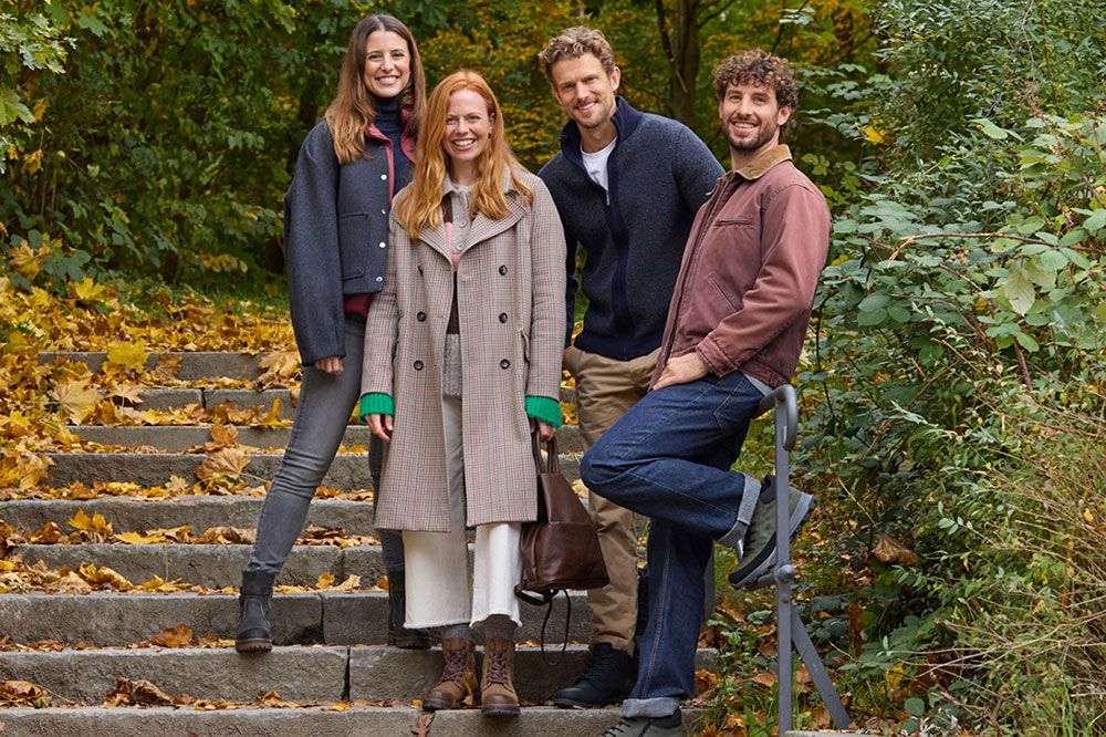 Vier Personen in Herbstkleidung stehen auf einer Treppe, umgeben von Laub und Bäumen, und lächeln in die Kamera.