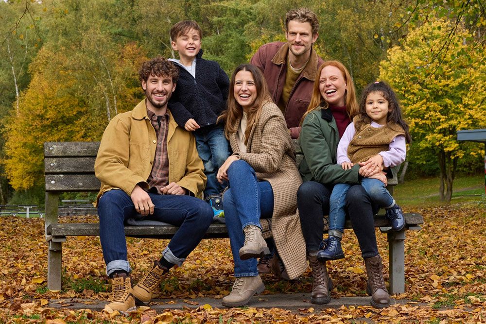 Eine Gruppe von sechs Personen, darunter zwei Kinder, sitzt auf einer Bank in einem Park, im Hintergrund sind Herbstblätter und bunte Bäume zu sehen.