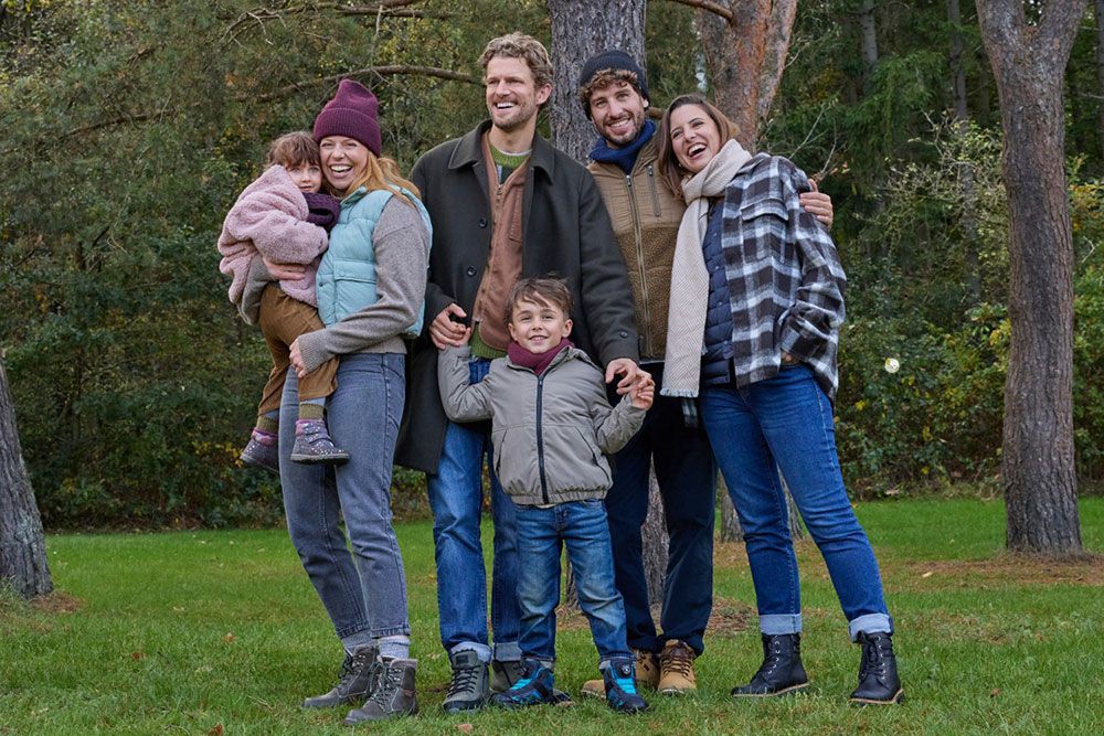 Eine Gruppe aus vier Erwachsenen und zwei Kindern steht lächelnd in einer grasbewachsenen, bewaldeten Gegend und trägt herbstliche Kleidung.