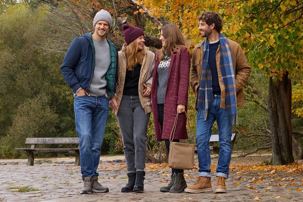 Vier Menschen in herbstlicher Kleidung stehen auf einem Kopfsteinpflasterweg, umgeben von bunten Herbstblättern, lächeln und genießen den Tag.