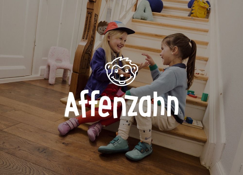 Zwei Kinder sitzen auf einer Holztreppe und spielen, beide lächeln. Das Logo der Marke Affenzahn ist im Vordergrund.