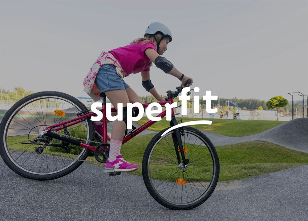 Mädchen mit Helm fährt auf einem rosa Fahrrad auf einem gepflasterten Weg in einem Park. Das Logo der Marke superfit überlagert das Bild.