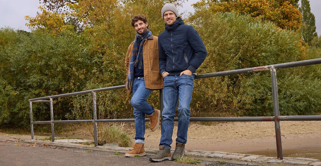 Zwei Männer in Herbstkleidung stehen an einem Geländer in einem Park, hinter ihnen sind üppiges grünes Laub und sandiger Boden zu sehen.