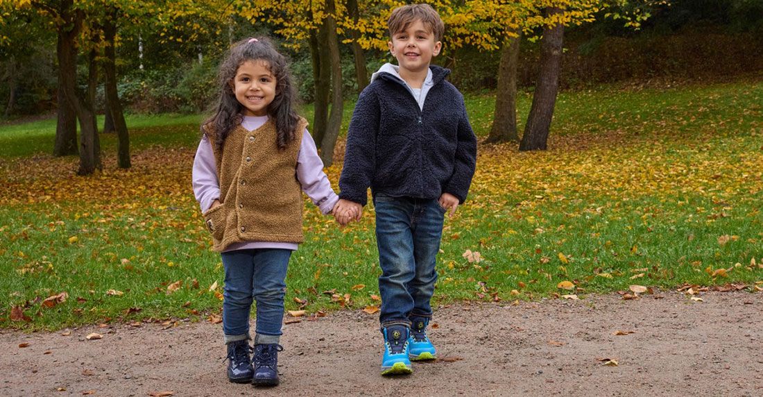 Zwei Kinder, die Händchen halten und lächeln, gehen im Herbst in einem Park spazieren, umgeben von Bäumen mit gelben Blättern.