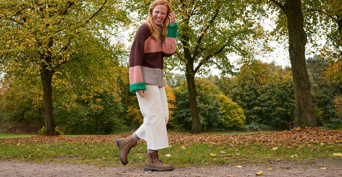 Eine Frau in einem Strickpullover, einer weißen Hose und braunen Steifeln lächelt in einem Park mit herbstlichen Bäumen und Laub.