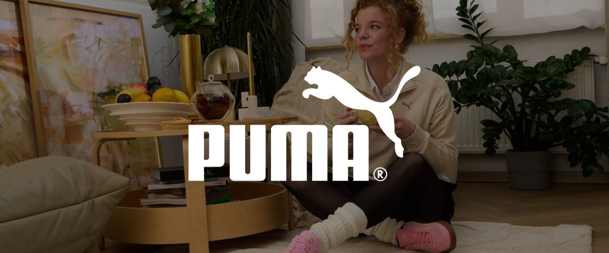 Eine Frau sitzt auf dem Boden, hält eine Tasse in der Hand, vor ihr steht ein Tisch mit Obst und Tee, umgeben von Pflanzen. Das Logo der Marke PUMA überlagert das Bild.