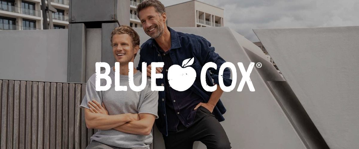 Zwei lächelnde Männer auf einem Dach, im Hintergrund moderne Gebäude. Das Logo der Marke BLUE COX überlagert das Bild.