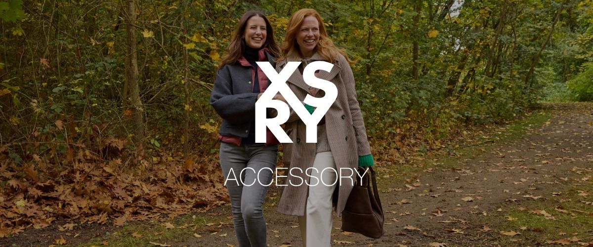 Zwei Frauen, die lächelnd durch einen herbstlichen Wald spazieren, Mäntel tragen und eine Tasche bei sich haben. Das Logo der Marke XSRY überlagert das Bild.