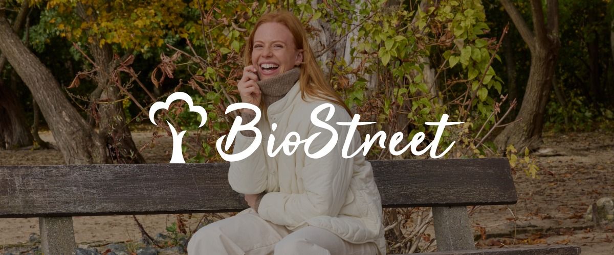 Eine Frau in einer weißen Jacke sitzt lächelnd auf einer Parkbank, umgeben von Herbstlaub. Das Logo der Marke Bio Street überlagert das Bild.