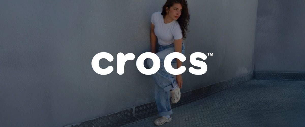 Eine Frau in einem weißen Oberteil und Jeans posiert vor einer Wand und trägt weiße Crocs. Das Logo der Marke Crocs überlagert das Bild.