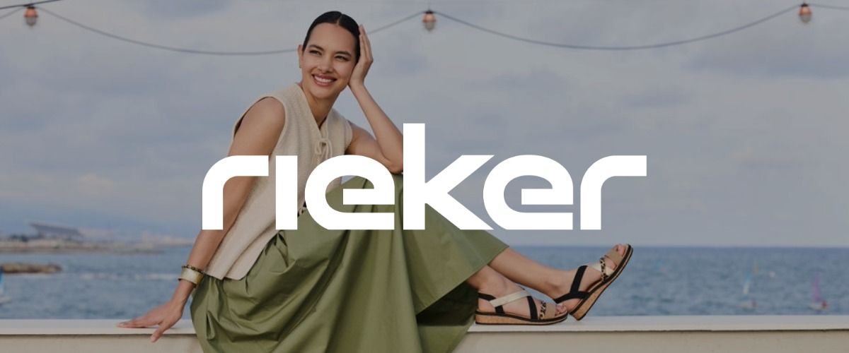 Eine Frau in einem hellen Oberteil und einem grünen Rock sitzt fröhlich auf einer Mauer am Meer. Das Logo der Marke Rieker überlagert das Bild.