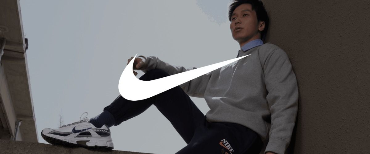 Ein Mann sitzt auf einer Mauer und trägt einen grauen Pullover, eine dunkle Jogginghose und weiße Nike-Sneaker. Das Logo der Marke Nike überlagert das Bild.