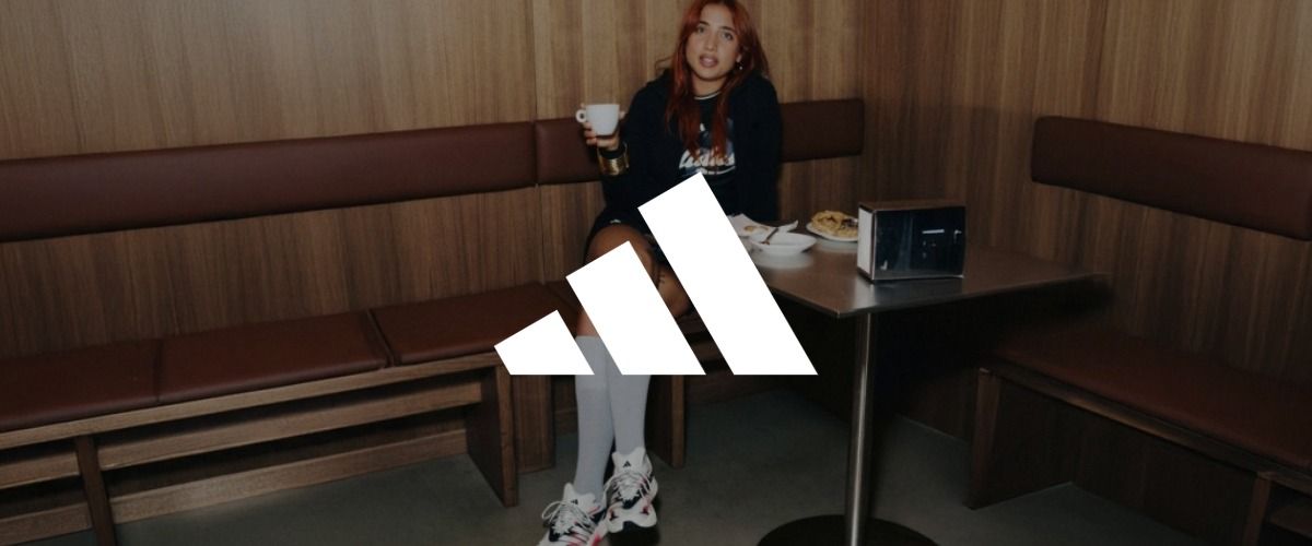 Eine Frau sitzt an einem kleinen Tisch mit Essen, trägt einen Kapuzenpulli und Turnschuhe und hält eine Tasse in der Hand. Das Logo der Marke adidas überlagert das Bild.
