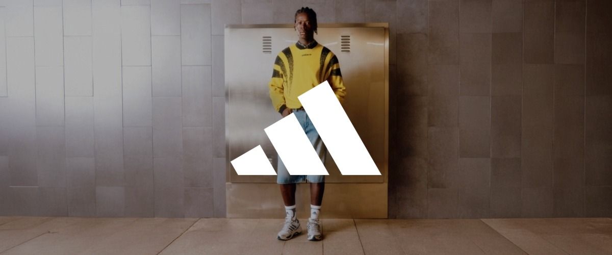 Ein Mann in einem gelben Pullover und Jeansshorts steht vor einer gefliesten Wand. Das Logo der Marke adidas überlagert das Bild.
