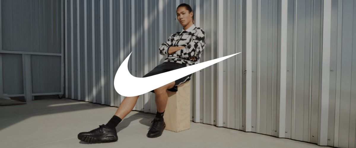 Ein Mann, der an einer Wellblechwand lehnt, eine karierte Jacke und Shorts trägt. Das Logo der Marke Nike überlagert das Bild.
