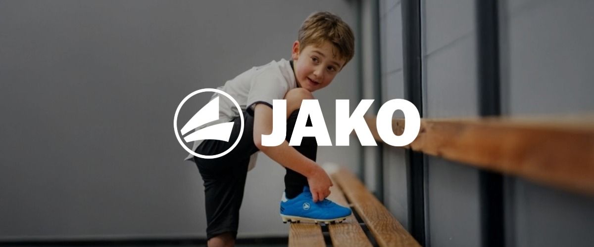 Ein kleiner Junge in Sportkleidung schnürt auf einer Bank in einer Umkleide seine hellblauen Stollenschuhe. Das Logo der Marke JAKO überlagert das Bild.